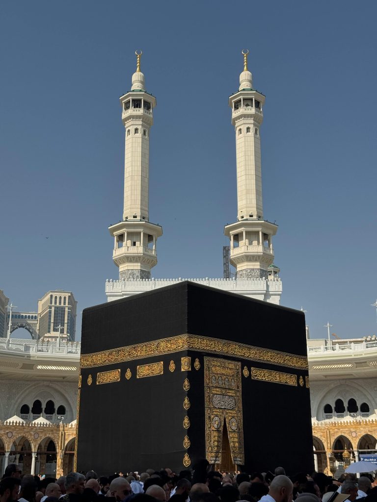 The Kaaba in Mecca, Saudi Arabia with a large crowd of pilgrims congregating under clear skies.
